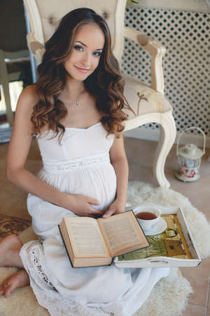 Pregnant woman reading a book sitting on the floor.Beautiful young pregnant woman, brunette with long curly hair and light makeup, sitting on the floor at home with an open book on her lapの写真素材