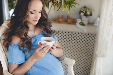 A young pregnant woman with a cup of tea.Beautiful young pregnant woman, brunette with long curly hair and light makeup, sitting on the floor at home with a cup of hot tea in his handsの写真素材