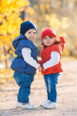 Little girls-girlfriends walk in the park.Two little girls happy friends playing together in the alley in the park on a background of yellow trees and bushes, dressed in jackets and knitted hats of red and blue on the legs wearing white shoesの写真素材
