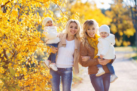 Happy moms, two blondes with long curly hair, wearing blue jeans and brown jackets, with their...