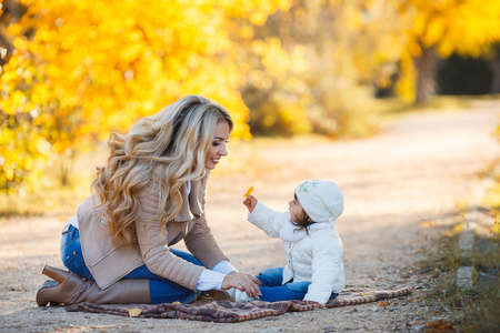 Mom and her little girl walk and have fun in a beautiful yellow in autumn city Park, her mother was blonde with long curly hair, in a beige jacket and blue jeans, a girl in a white jacket and a white cap, happy familyの写真素材