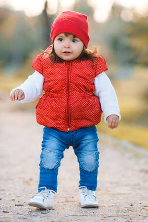 Little girl in yellow autumn park.Funny little girl in a red hat, red jacket, blue jeans, with big brown eyes, walks the walk in autumn Park with yellow trees and bushesの写真素材
