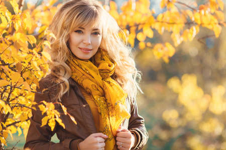 Beautiful woman in autumn park. Beautiful young blonde, woman with long curly hair, brown jacket, posing for a photograph on the background of shrubs and trees with bright yellow leaves in a city park on a sunny day.の写真素材