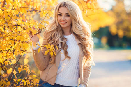 Beautiful woman in autumn park. Beautiful young blonde, woman with long curly hair, brown jacket, posing for a photograph on the background of shrubs and trees with bright yellow leaves in a city park on a sunny day.の写真素材