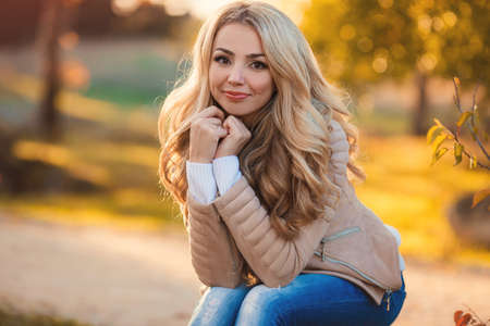 Beautiful woman in autumn park. Beautiful young blonde, woman with long curly hair, brown jacket, posing for a photograph on the background of shrubs and trees with bright yellow leaves in a city park on a sunny day.の写真素材