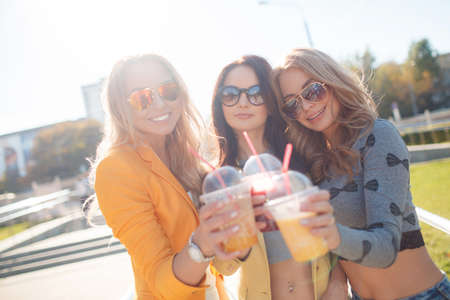 Three girls are walking in the Park in summer and drink fruit juice.Three young beautiful women, two blondes and a brunette, go for a walk together in the Park in the summertime, hands holding plastic cups, drink fruit juices and wear sun glassesの写真素材