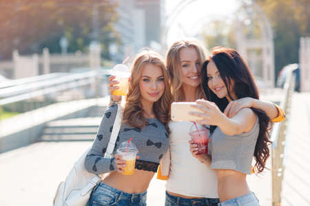 Three beautiful women take off their selfie on smartphone.Three beautiful young women, two blondes and a brunette, are walking together in the Park in summer time, drink fruit juices, smile and take a selfie on your smartphone