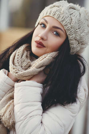 Autumn portrait of a beautiful young woman, brunette with long straight hair and gray eyes, poses for photographer in the fresh air in autumn, dressed in a white jacket, a beige knitted hat and scarf, beautiful makeupの写真素材