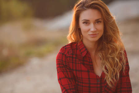 Woman portrait outdoor in sunset light.Cute young woman with long curly red hair and brown eyes, wearing dark jeans and a red plaid shirt, pretty smile, posing in the setting sun, sitting cross-legged outdoors in summerの写真素材