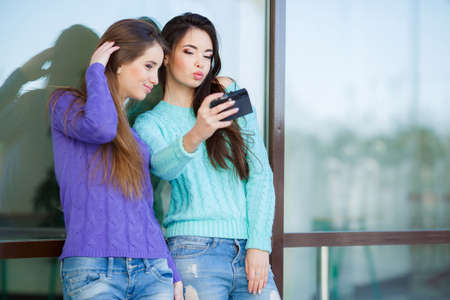 Two cute girl, brunette with long straight hair, both dressed in blue jeans and knitted sweaters, stand near the large glass windows with a retro camera in hand and take pictures in his hometownの写真素材
