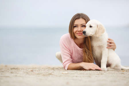 Beautiful young female model with sweet smile, brunette with long straight hair and gray-green eyes, light makeup, in a pink knitted sweater, walks along the beach with his friend-a puppy Golden Retrieverの写真素材