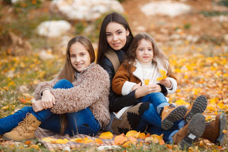 Three beautiful girls of different ages, eastern appearance, brunette with brown eyes and long hair, cute smile, the three happy sisters relax and have fun together outdoors in yellow autumn parkの写真素材