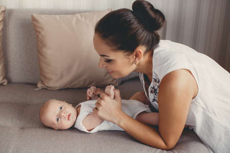 A young mother, brunette, hair pulled back, white sleeveless blouse, spending time together with their newborn son at home in the bedroom, talking and playing with the boy, dressed in a white t-shirt and white pantiesの写真素材