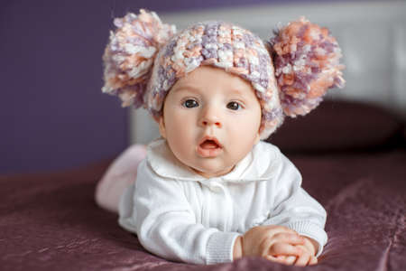 Portrait of a happy baby crawling on the bed.Little girl 3 months, with brown eyes, dressed in a white blouse and pink romper, knitted cap with a large bubo, stares at the photographer, raising his head lying on a purple blanketの写真素材