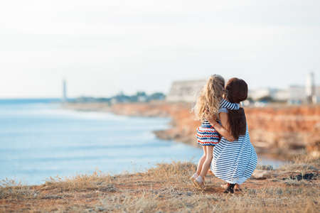 A slim young woman, a brunette, dressed in a short white dress with dark blue stripes, spends time together with his little cute daughter on beach near the ocean, a girl with beautiful blonde curly hair, dressed in a striped t-shirt and skirt , summerの写真素材
