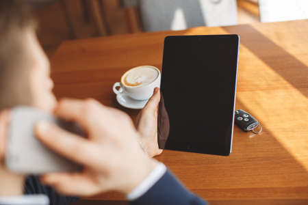 A handsome young man, dark hair with short hair, dressed in a white shirt and a dark blue jacket, holding a tablet computer and to call on his cell phone, wearing a watch on his left wrist, sitting at a table in a cafeの写真素材