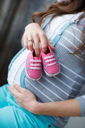 Beautiful young pregnant woman, a brunette with long hair, dressed in a gray sweater and blue pants, right hand wedding ring, holding pink booties for the baby, putting them on his big bare bellyの写真素材