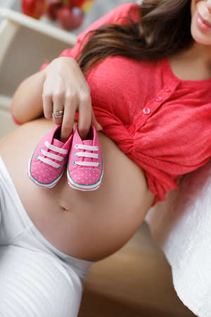 Beautiful young pregnant woman, brunette with long thick hair, wearing a red shirt and white pants, right hand wedding ring, holding pink booties for the baby, putting them on his big bare bellyの写真素材