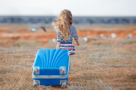 The little girl with a big blue suitcase.A little girl with long blonde curly hair, in a striped shirt and knitted skirt with a large blue suitcase on wheels is on the field with dry yellow grass, the view from the backの写真素材