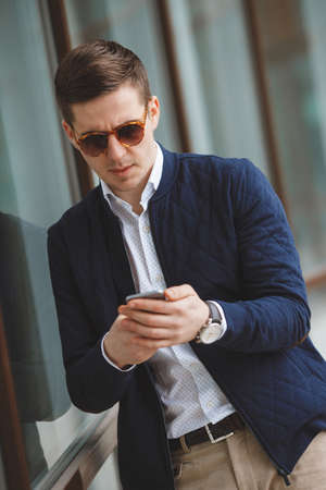 Business young man, dark short hair, dressed in a white shirt, dark blue jacket and light brown pants with a waist belt, wearing a wrist watch, standing outdoors near a glass office building, talking on a cell phoneの写真素材