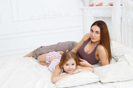 Young beautiful woman brunette with long hair and brown eyes, together with his daughter, a girl of 6 years, brown hair, brown eyes, dressed in a t-shirt and panties, to spend time together in the bedroom, lying on a white bedの写真素材