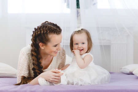 Happy family, young beautiful woman with dark hair, in a braid, gray eyes, dressed in a white lace dress, playing with his little daughter, girl - a brunette with short hair, in the bedroom on the bed with purple blanketの写真素材