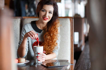 A red-haired woman with a Cup of coffee in the cafe.A bright, beautiful woman with curly red hair, red lipstick, beautiful makeup, red nail Polish, sitting alone at a table in a cafe with a Cup of coffee latte and vanilla biscuitsの写真素材