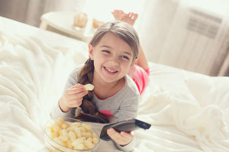 The pretty little girl of 6 years, the brunette with the long hair braided to the spit is dressed in a gray t-shirt and pink striped trousers, spends the time of the house, lies on a white bed near a window with the control panel the TV in one hand, eats の写真素材