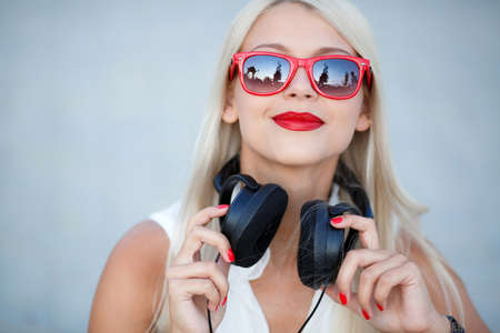 Beautiful young woman with long blonde hair and grey eyes, red lipstick, large black headphones, dressed in a white sleeveless shirt, wears sun glasses in the red frame, enjoy the music, posing on a blue backgroundの写真素材