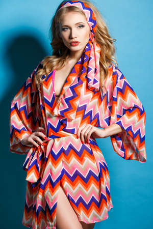 Beautiful sexy blonde model with lovely wavy long hair and a beautiful figure, pink lipstick and light makeup, red nail Polish, posing in bright, colorful, short robe on a blue background in the Studioの写真素材