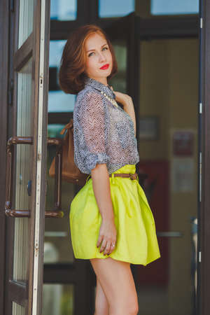 The beautiful woman with a nutbrown hair and a stylish hairdress, gray eyes and the red lipstick dressed in a skirt of lemon color and a gray blouse wears a white necklace, red nail varnish, poses with a brown bag at an entrance to a supermarketの写真素材