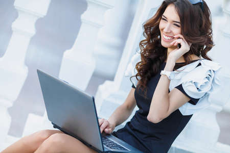 Cute business woman brunette with long curly hair, dressed in a black dress with white sleeves, sitting on the steps near the white railing, works with laptop and talking on mobile phone outdoorsの写真素材