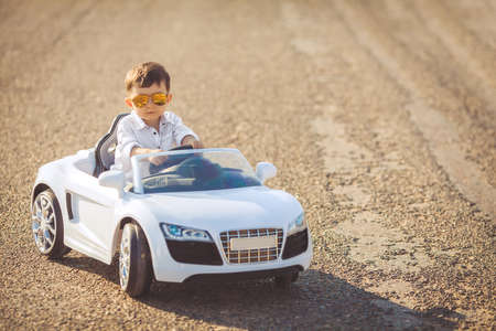 The young driver, a boy with brunette short hair, mirrored sun glasses yellow, in white shirt posing on a mountain road against the sea and clear sky, sitting in a posh toy car white color in the fresh summer airの写真素材