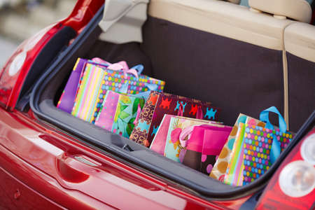 Open the trunk of the car with bags of purchases.The red car is a hatchback, the seats beige color, stands alone near the entrance to the shopping centre with an open trunk lid, filled with colored paper bags for shopping aheadの写真素材