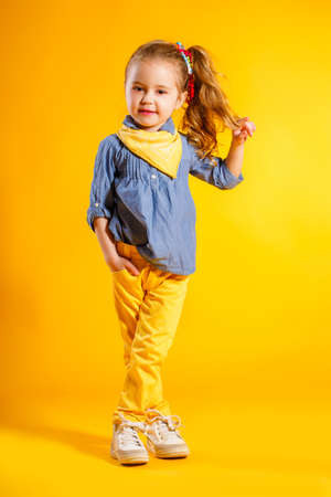 Portrait of a cute small girl, brunette with brown eyes and long curly hair, tied with a ribbon, a shirt of blue-gray and yellow handkerchief around his neck, yellow pants, posing in Studio on a bright yellow backgroundの写真素材