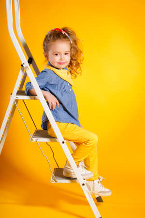 Portrait of a cute small girl, brunette with brown eyes and long curly hair, tied with a ribbon, a shirt of blue-gray and yellow handkerchief around his neck, yellow pants, posing in Studio on a bright yellow background with ladderの写真素材