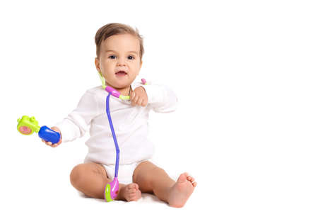 Portrait of a curious little boy, brunette with brown eyes and short hair, dressed in a white shirt and a white diaper, barefoot posing in Studio, sitting on a white background, smiling mouth open, plays with a toy stethoscopeの写真素材