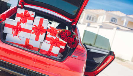 Gift boxes in a luggage carrier of the red car.White boxes with gifts tied with red ribbons with large red bows and a huge bouquet of red roses, neatly stacked in the open trunk of the big red car, the concept of celebration and loveの写真素材