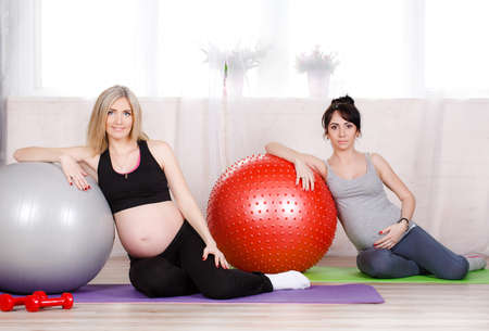 Two happy pregnant women, brunette and blonde, dressed in sportswear, execute a complex of exercises for pregnant women with large gymnastic balls, grey and red, training is carried out in a bright room fitness centreの写真素材