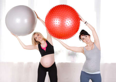 Two happy pregnant women, brunette and blonde, dressed in sportswear, execute a complex of exercises...