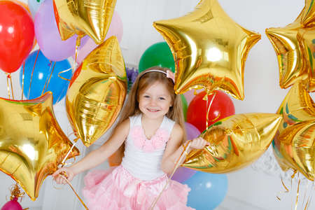 Beautiful child, redhead girl with long straight hair and grey eyes, with a pink bow on her head, dressed in pink and white dress on a white background with multi-colored balloons and gold stars to your fourth birthdayの写真素材