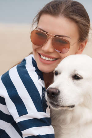 Portrait of young beautiful woman in dark glasses, brunette with beautiful smile, wearing a striped t-shirt and blue jeans, sitting on a sandy beach against the blue of the ocean, hugging a beloved friend - a dog breed Golden Retrieverの写真素材