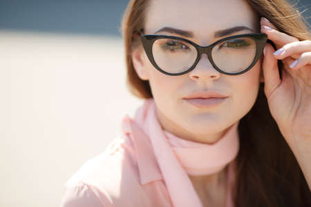 Young beautiful woman brunette with long straight hair and brown eyes, dressed in a pink blouse and pink scarf around his neck, wears black rimmed glasses, posing for the photographer in the summer outdoorsの写真素材