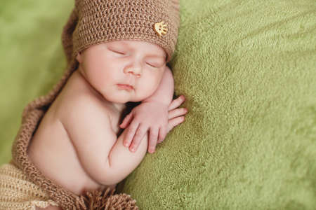 Peaceful sleep of a newborn baby. Newborn baby in brown knitted hat sleeping sweetly on a soft green blanket handles himself under his cheekの写真素材