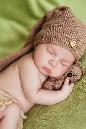 Peaceful sleep of a newborn baby. Happy newborn baby in knitted brown hat, sleeping sweetly on a soft green bed, putting the pen under his cheek and hugging toy brown bearの写真素材