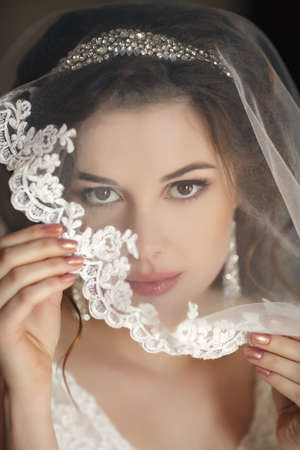 The young beautiful woman, the brunette with a wedding hairdress and a professional make-up, in ears expensive earrings, is dressed in a white wedding dress and a transparent white veil, waits for the groom in a hotel roomの写真素材