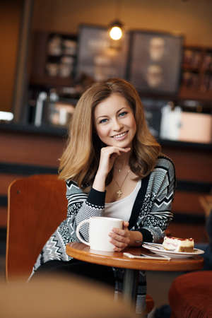 Young beautiful woman with long, blond, thick hair and gray eyes, on the neck is a gold cross, wearing a gray jacket with a black pattern and a white t-shirt, sitting alone at a table in a cozy cafe with a big white cup of hot teaの写真素材