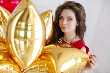 Young beautiful brunette woman with long curly hair and grey eyes, dressed in a red sleeveless dress, round neck wears a white necklace, a portrait, on light gray background in the Studio with a bunch of gold balloons - of pillows on your birthdayの写真素材