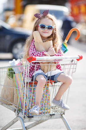 Little girl, with thick blonde long hair, dressed in a plaid shirt and denim shorts, on his head a black-and-red bow, wears sun glasses in blue frame, sitting in the shopping cart on wheels near a large supermarketの写真素材