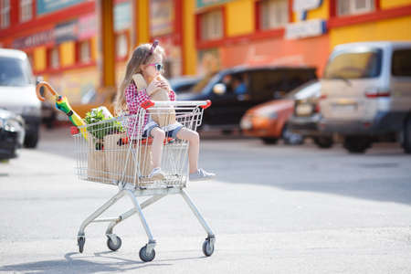 Little girl, with thick blonde long hair, dressed in a plaid shirt and denim shorts, on his head a black-and-red bow, wears sun glasses in blue frame, sitting in the shopping cart on wheels near a large supermarketの写真素材